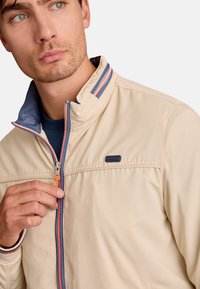 Veste beige zippée avec un col montant, ornée d'accents bleus et rouges, en tissu texturé, avec un logo près de la fermeture éclair, portée partiellement zippée.