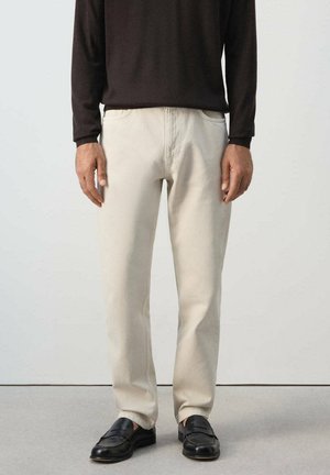 Mann trägt beige gerade geschnittene Hose, schwarze Loafers und ein dunkelbraunes langärmeliges Hemd und steht vor einem schlichten Hintergrund.