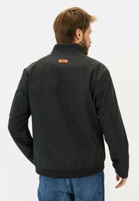 Sort jakke med glat tekstur, stående krave og et brunt lædermærke i midten af ryggen. Har ribbede manchetter og kant.