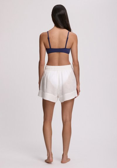 Γυναίκα που στέκεται ξυπόλυτη πλάτη, φοράει σκούρο μπλε crop top και λευκά φαρδιά σορτς σε μονόχρωμο φόντο.