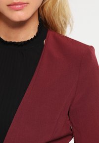 Gros plan sur une personne portant un haut noir à col montant avec volants et un blazer bordeaux, montrant les lèvres, le cou et l'épaule.