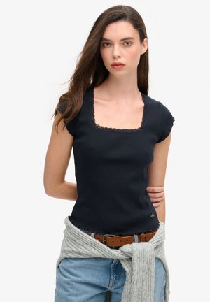 Top de punto acanalado en color navy con mangas cortas y escote cuadrado con detalles de encaje. Combinado con jeans casuales y un chal de punto gris alrededor de la cintura.