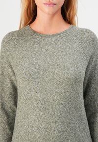 Woman wearing a light olive-green knitted crew neck sweater, showing upper torso and part of face with natural lip gloss.