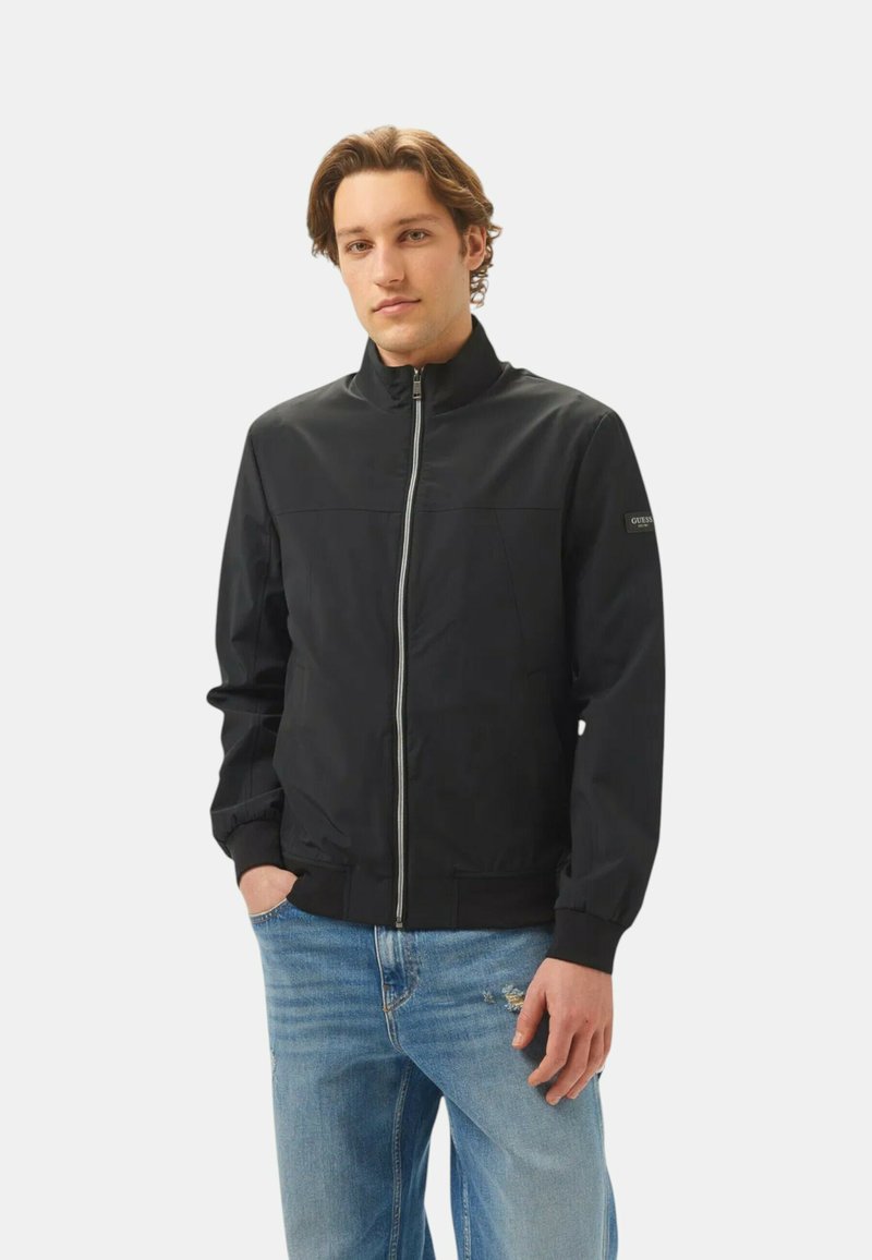 Giacca nera con zip e colletto alto, polsini e orlo elastici. Tessuto liscio con un design minimalista. Indossata sopra jeans chiari strappati.