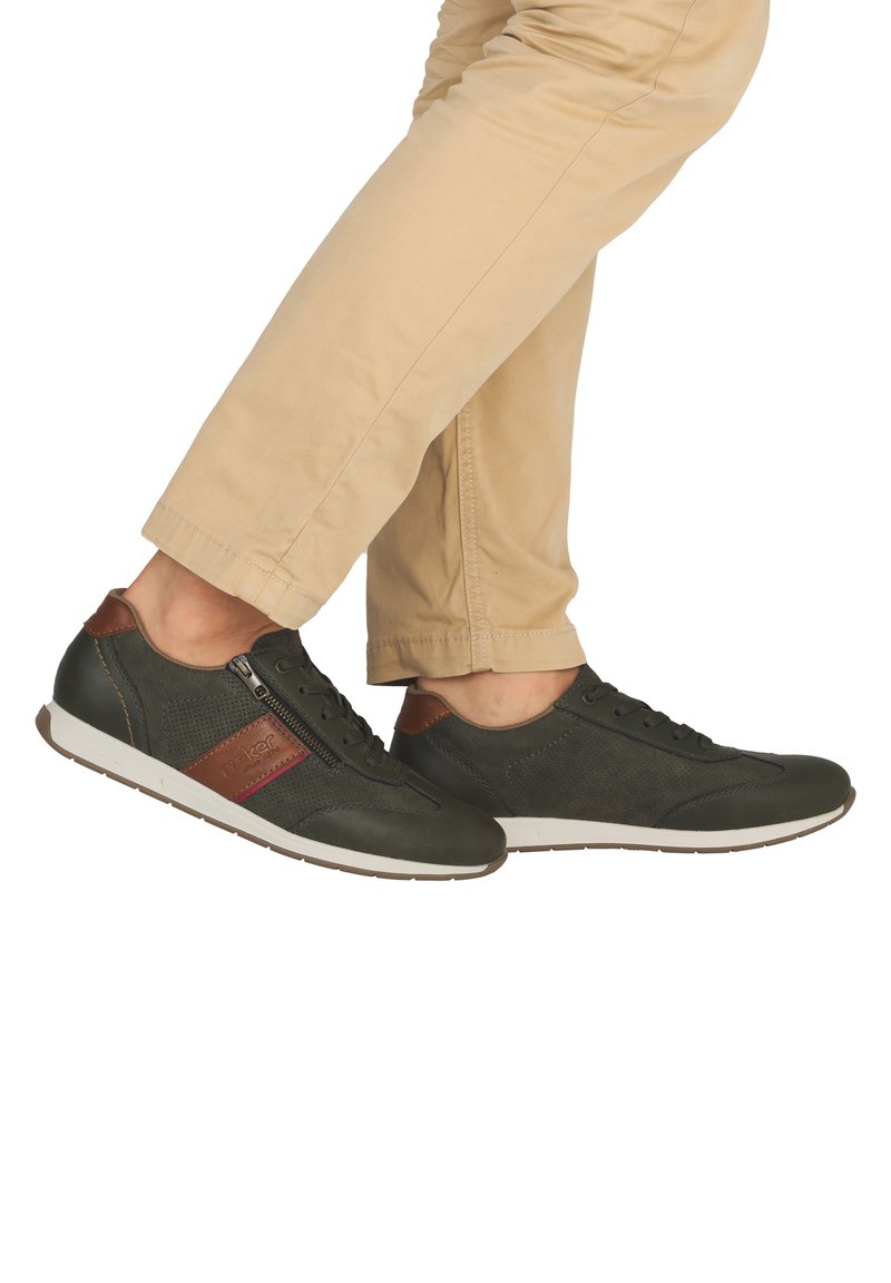 Personne portant un pantalon beige et des chaussures décontractées vert foncé avec des accents marron et une fermeture éclair latérale, debout sur un fond blanc.