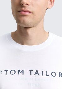 Detailaufnahme einer Person, die ein weißes T-Shirt trägt, auf dem in großen, verblassten schwarzen Buchstaben "TOM TAILOR" über die Brust gedruckt ist.