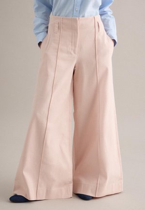Pantaloni rosa chiaro a gamba larga e vita alta, indossati con una camicia azzurro chiaro infilata dentro, mani nelle tasche e scarpe scure visibili.