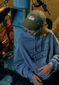Khakifarbene Baseballkappe mit „Tigers“-Stickerei, grauer Kapuzenpullover mit „Stranger Things“-Logo, urbanes Umfeld, Fahrrad im Hintergrund.