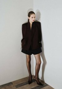 Blazer burdeos oscuro con hombros pronunciados, llevado sobre una blusa clara y pantalones cortos negros. Medias negras transparentes y tacones afilados completan el conjunto.