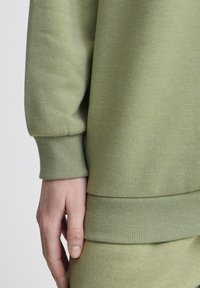 Maglione verde chiaro con polsini e orlo a costine, realizzato in materiale morbido. Ingrandimento di una mano appoggiata su un dettaglio del tessuto.