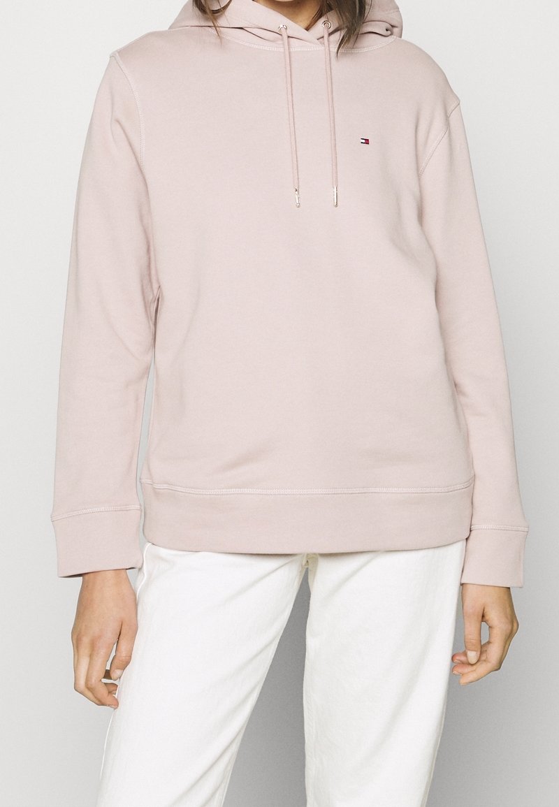 Femme portant un sweat à capuche rose clair avec cordons à l'avant et un pantalon blanc, debout devant un fond uni.