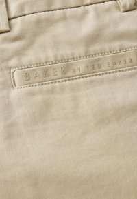 Béžová látka so zašitou štítkom s nápisom "BAKER BY TED BAKER" pri opaskovom očku na páre nohavíc alebo kraťasov.