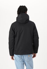 Chaqueta negra con capucha de textura suave, con líneas sencillas y sin herrajes visibles. Llevada sobre unos vaqueros azul claro.