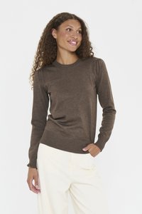 Femme aux cheveux frisés portant un pull long à manches marron et un pantalon crème, se tenant avec une main dans la poche contre un fond blanc.