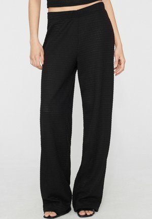 Vrouw draagt hoge taille, wijde zwarte textuurbroek en zwarte open-toe schoenen, staand voor een effen achtergrond.