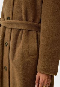 Bruine wollen jas met een zachte textuur, een knoopsluiting aan de voorkant, zijzakken en een bijpassende stoffen riem voor aanpassing.