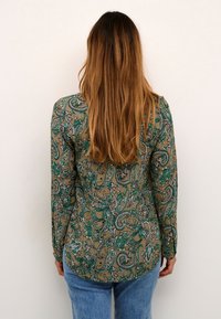 Chemise à manches longues avec un imprimé paisley vert sur fond marron, dotée d'un ourlet arrondi et de poignets boutonnés, associée à un jean bleu.