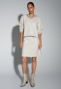 Pullover à capuche beige et jupe assortie, toutes deux en tissu doux à la texture lisse. Le modèle porte également des bottines beiges brillantes à talon bloc.