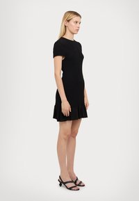 Melnā pieguļošā kleita ar īsām piedurknēm un ruffled apakšmalu. Uzbūvēta no teksturēta auduma, tai ir vienkāršs apaļš izgriezums. Nostaigāta ar melniem čību sandaliem.