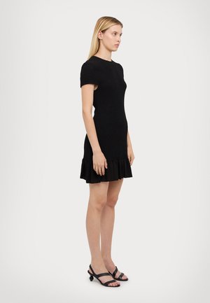 Black fitted dress with short sleeves and a ruffled hem. Made of textured fabric, it features a simple round neckline. Worn with black sandals.