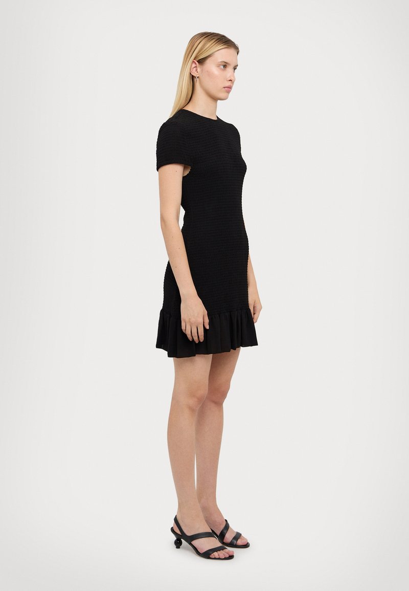 Melnā pieguļošā kleita ar īsām piedurknēm un ruffled apakšmalu. Uzbūvēta no teksturēta auduma, tai ir vienkāršs apaļš izgriezums. Nostaigāta ar melniem čību sandaliem.