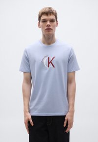 Ανοιχτό μπλε βαμβακερό T-shirt με κυκλικό λογότυπο Calvin Klein σε κόκκινο και κρεμ. Κοντά μανίκια, λαιμόκοψη στρογγυλή και κανονική εφαρμογή.