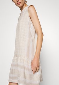 Helles beige ärmelloses Kleid mit strukturiertem Muster, gerafftem Saum und subtilen Streifen in verschiedenen Farbtönen, aus weichem Stoff.