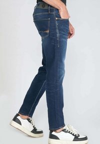 Jeans en denim bleu avec une coupe droite, texture délavée et détails de couture subtils. Portés avec des baskets blanches et noires.