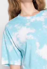 Koszula z krótkim rękawem w kolorze błękitnym, ombre, charakteryzująca się miękką fakturą i białymi wzorami przypominającymi chmury na tkaninie. Okrągły dekolt.