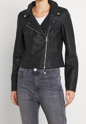 Personne portant une veste de motard en similicuir noire avec une fermeture éclair argentée et un jean taille haute gris foncé sur un fond uni.