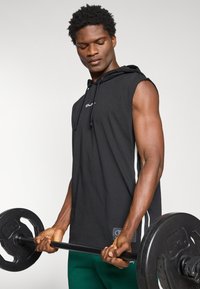 Man lifting barbell with weight plates, wearing sleeveless black hoodie and green pants against plain background.