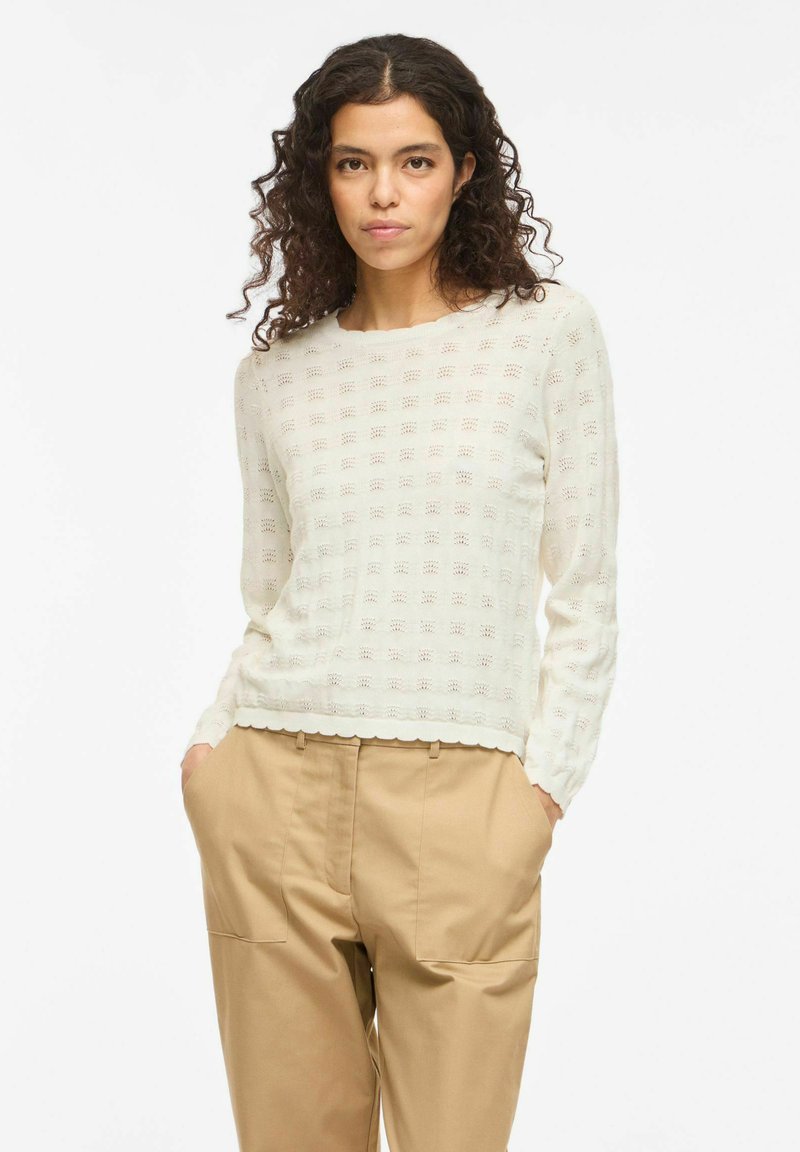 Femme aux cheveux bouclés et foncés portant un haut blanc texturé à manches longues et un pantalon beige, les mains dans les poches, sur fond blanc.