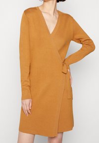 Vestito avvolgente giallo senape con scollatura a V, maniche lunghe e dettaglio di legatura laterale. Tessuto a maglia testurizzato con una finitura liscia.