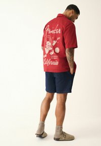 Homme portant une chemise à manches courtes rouge avec un graphique de guitare Fender et le texte "California", un short bleu marine, des chaussettes beiges et des chaussures à enfiler, les mains dans les poches.