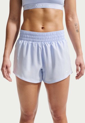 Nainen pukeutuneena vaaleansinisiin Nike urheilushortseihin ja vastaavaan urheiluliiviin, näyttää keskivartalon ja reisien yläosan yksiväristä taustaa vasten.