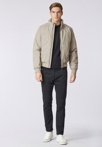 Giubbotto bomber beige con colletto alto, chiusura con zip, polsini elasticizzati e vita aderente, abbinato a jeans neri e sneakers bianche.