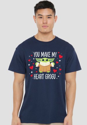Young man wearing navy blue t-shirt with cartoon Grogu and red hearts, text "You make my heart Grogu" on front.