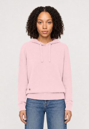 Woman with curly hair wearing a light pink hoodie with drawstrings and blue jeans, standing against a plain light background.
