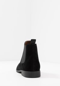 Musta mokkanahkainen nilkkuri, jossa on joustavat sivupaneelit, litteä pohja ja vetonauha kantapäässä. Sileä pinta, jossa on minimaaliset ompeluyksityiskohdat.