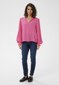 Blouse fuchsia à manches longues avec un col en V, poignets élastiques, coupe ample, associée à un jean skinny bleu foncé et des mocassins noirs.