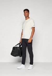 T-shirt bege de algodão com logótipo "Champion", calças justas pretas, ténis brancos e uma mala desportiva preta. Modelo em pose neutra.