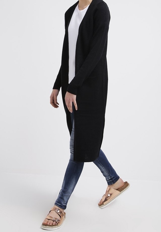 Personne marchant portant un long cardigan noir, un haut blanc, un jean skinny bleu et des sandales métalliques couleur or rose avec des brides à la cheville.
