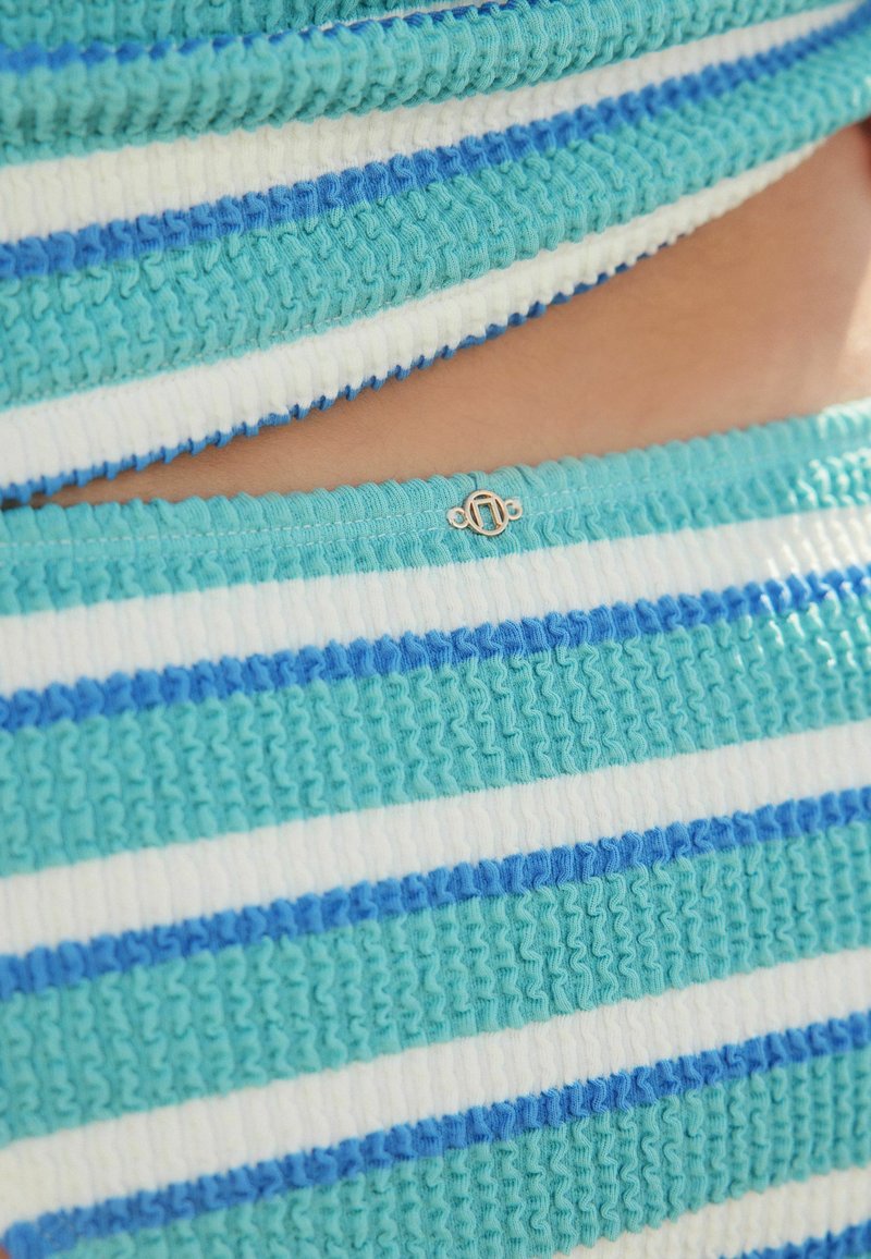 Dzianina w niebiesko-białe paski o teksturowanym wykończeniu i małym złotym akcentem na dole. Charakteryzuje się marszczonymi krawędziami i paskami o różnej grubości.