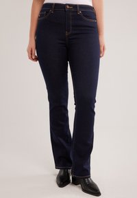 Žena odjevena u tamnoplave traperice bootcut kroja i crne gležnjače s potpeticom, stoji nasuprot jednostavnoj svijetloj pozadini.