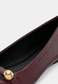 Μοκασίνι από δέρμα μπορντό με στρογγυλεμένη μύτη, που διαθέτει χρυσό διακοσμητικό κουμπί και εσωτερική επένδυση σε μαύρο χρώμα.