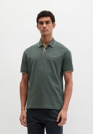 Junger Mann stehend, trägt ein dunkelgrünes Polohemd mit kurzen Ärmeln, kontrastierendem Kragenbesatz und dunkle Hose, neutraler Hintergrund.