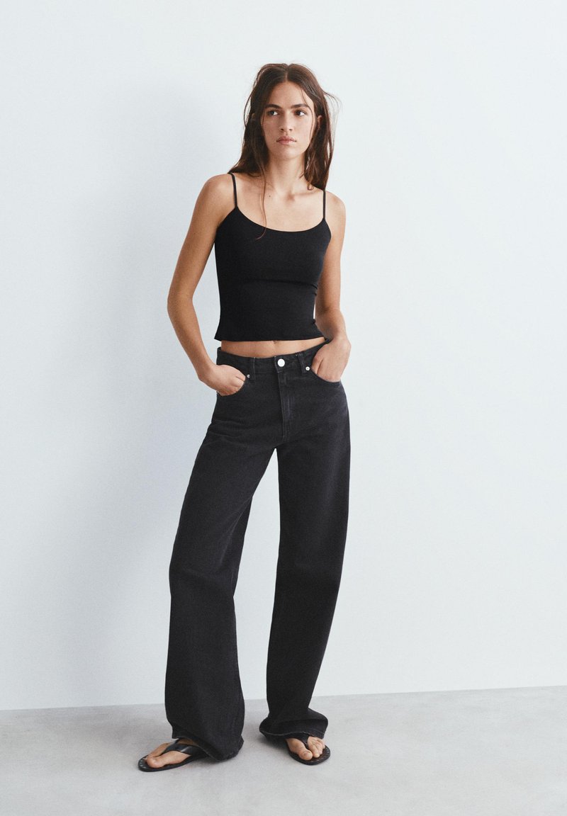 Jeune femme debout, les mains dans les poches, portant un top noir à fines bretelles, un jean noir à jambes larges et des tongs noires devant un mur uni.