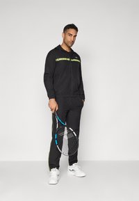Czarny sportowy dres z zielonymi detalami, z zamkiem błyskawicznym, kurtka oraz pasujące dresy. Postać trzyma rakietę tenisową i ma na sobie białe sneakersy.