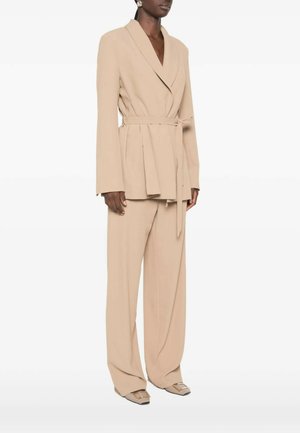 Vrouw draagt een beige blazer met riem, wijde broekspijpen en beige puntige schoenen met vierkante versieringen.
