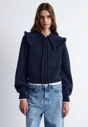 Marineblau gestrickte Strickjacke mit Rüschenkragen, Knopfverschluss und langen Puffärmeln; getragen über hellblauen Jeans. Strukturierter Stoff.